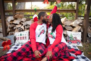 Couple in buffalo plaid and Christmas shirts relaxes against stacked firewood background.