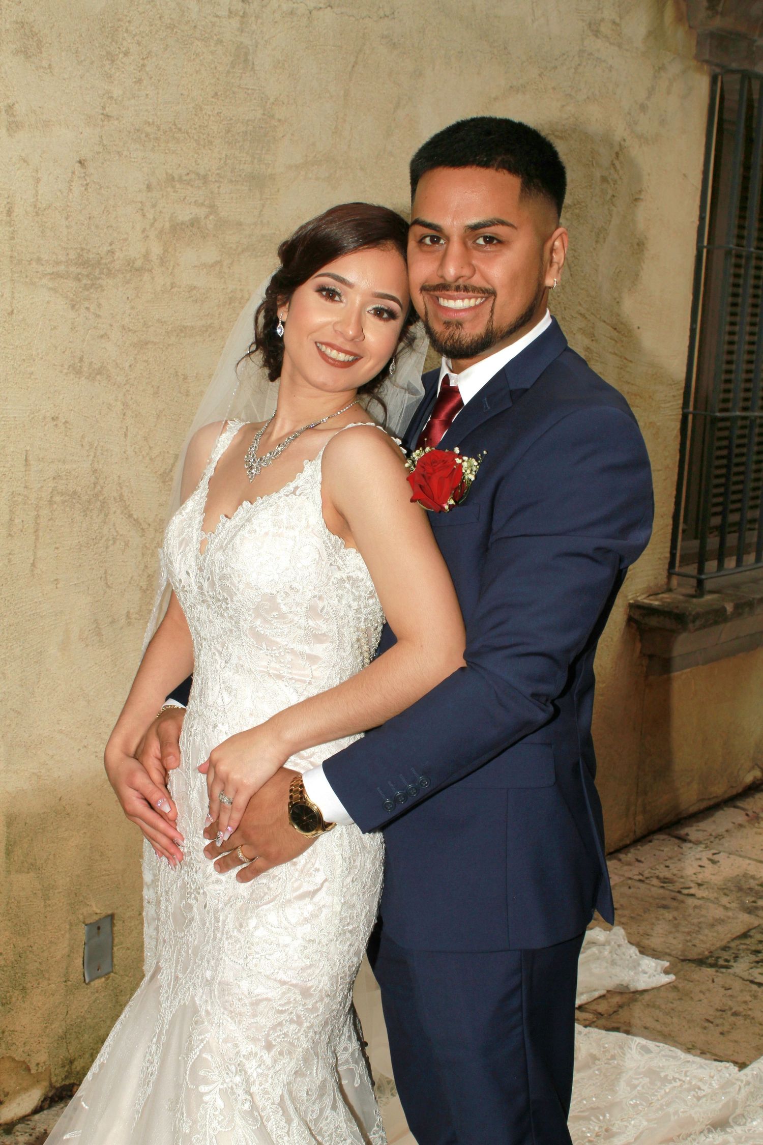 Newlyweds pose against a textured beige wall in formal wedding attire.