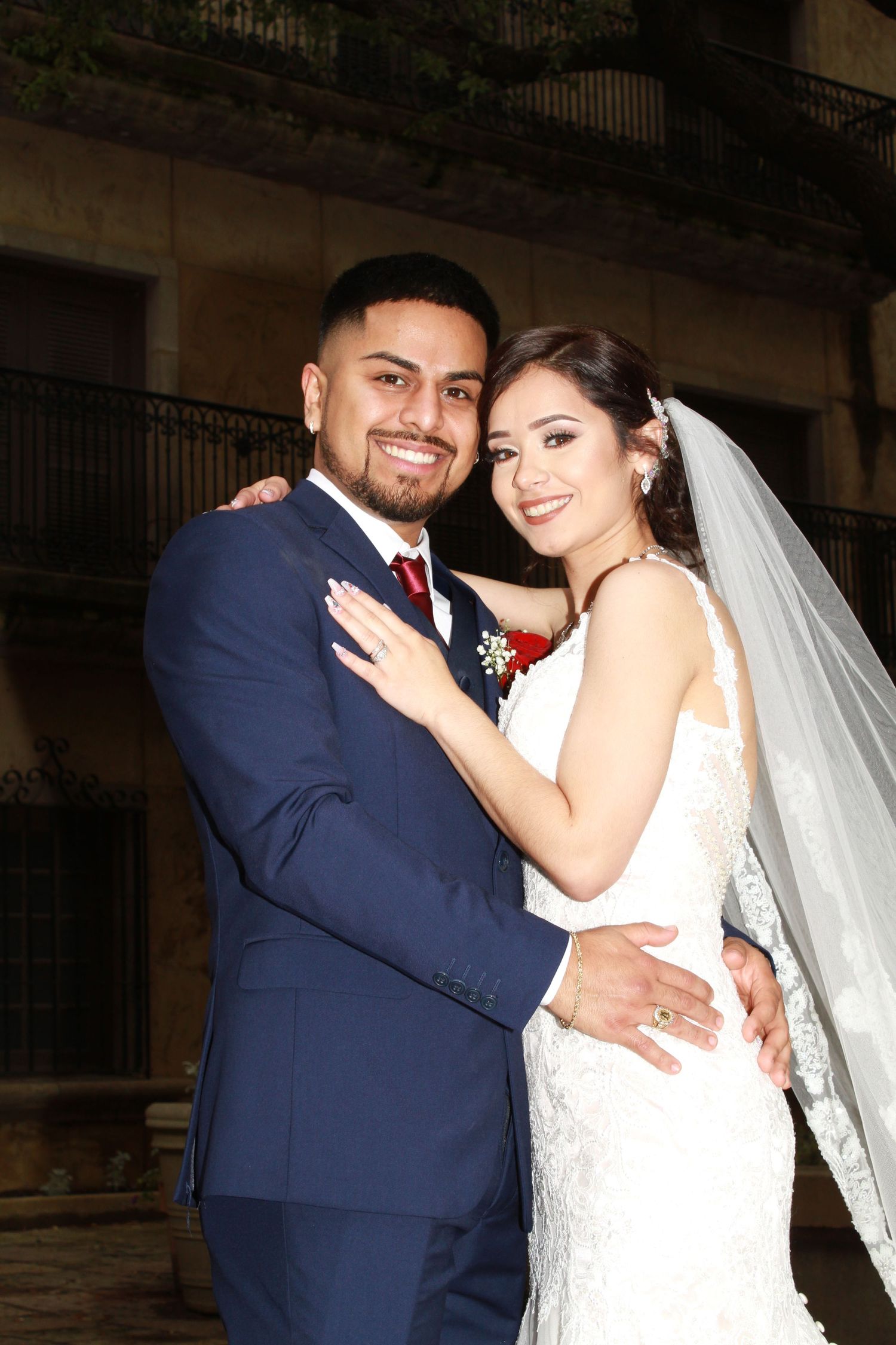 A couple in formal wedding attire embrace against a historic building backdrop.