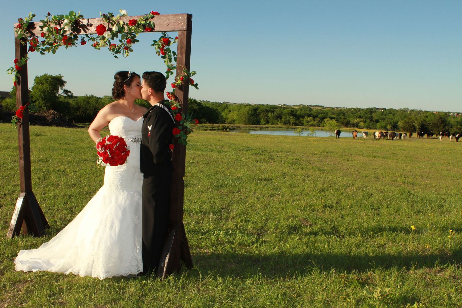 Wedding ceremony setup in a scenic rural field with wooden arch decorated with red flowers.