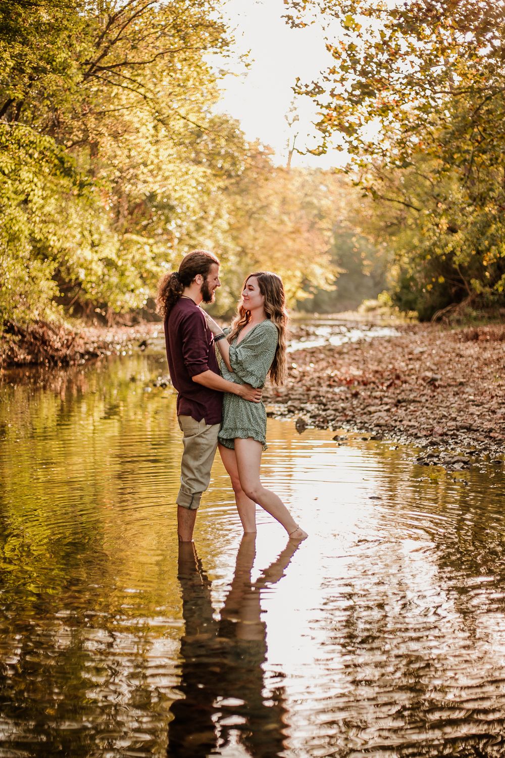 A romantic couple stands together in shallow creek water surrounded by autumn foliage at sunset.