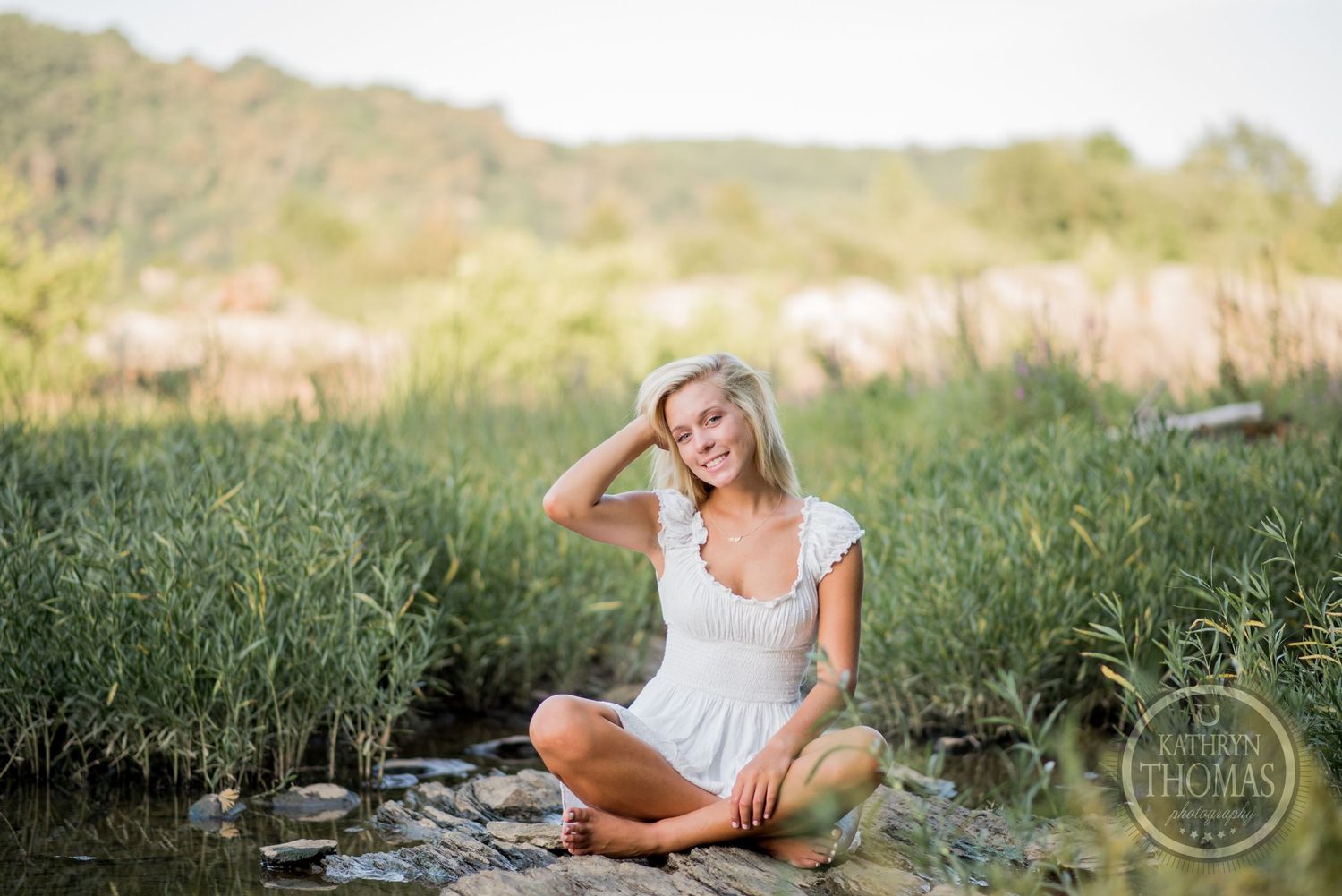 Outdoor portrait by water shows someone in white dress sitting cross-legged in tall grass.