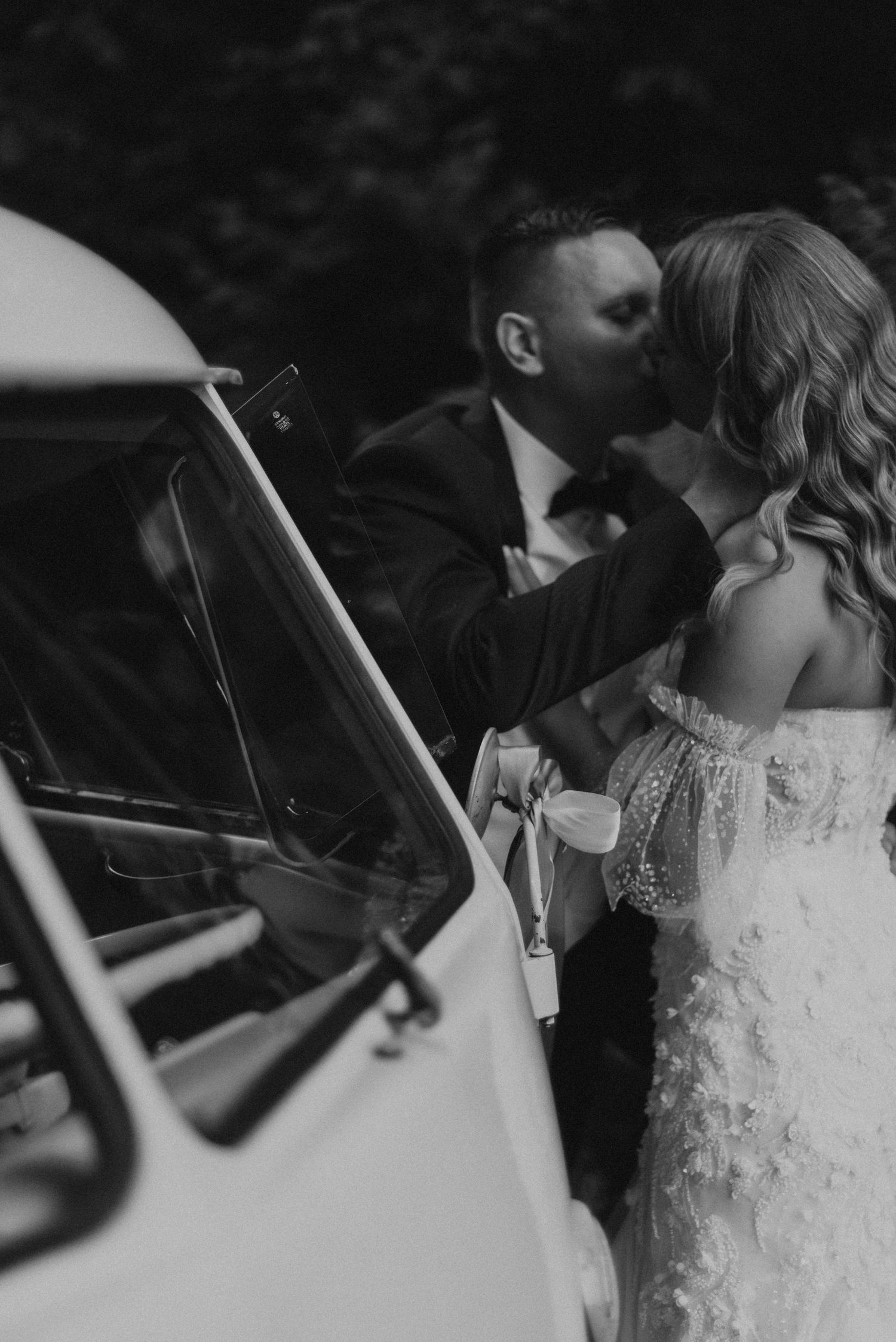 A romantic black and white photo of a couple by a vintage car on their wedding day.