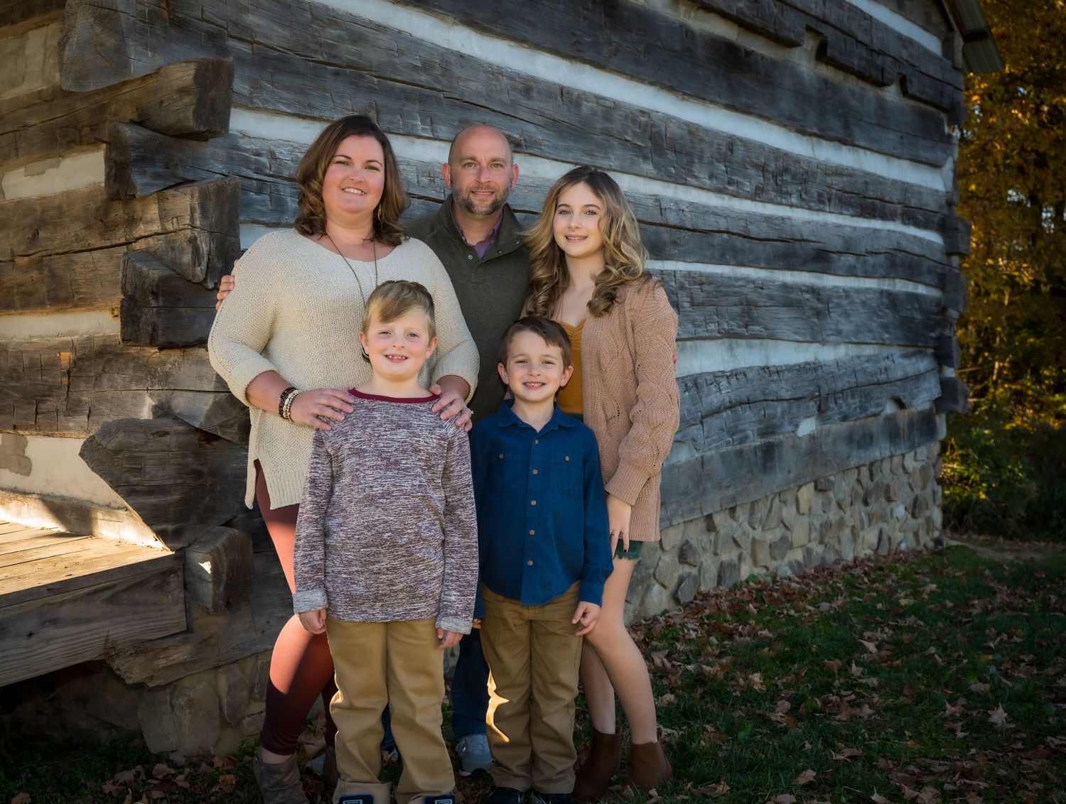A family of five poses together against a rustic log cabin wall during an outdoor portrait session.