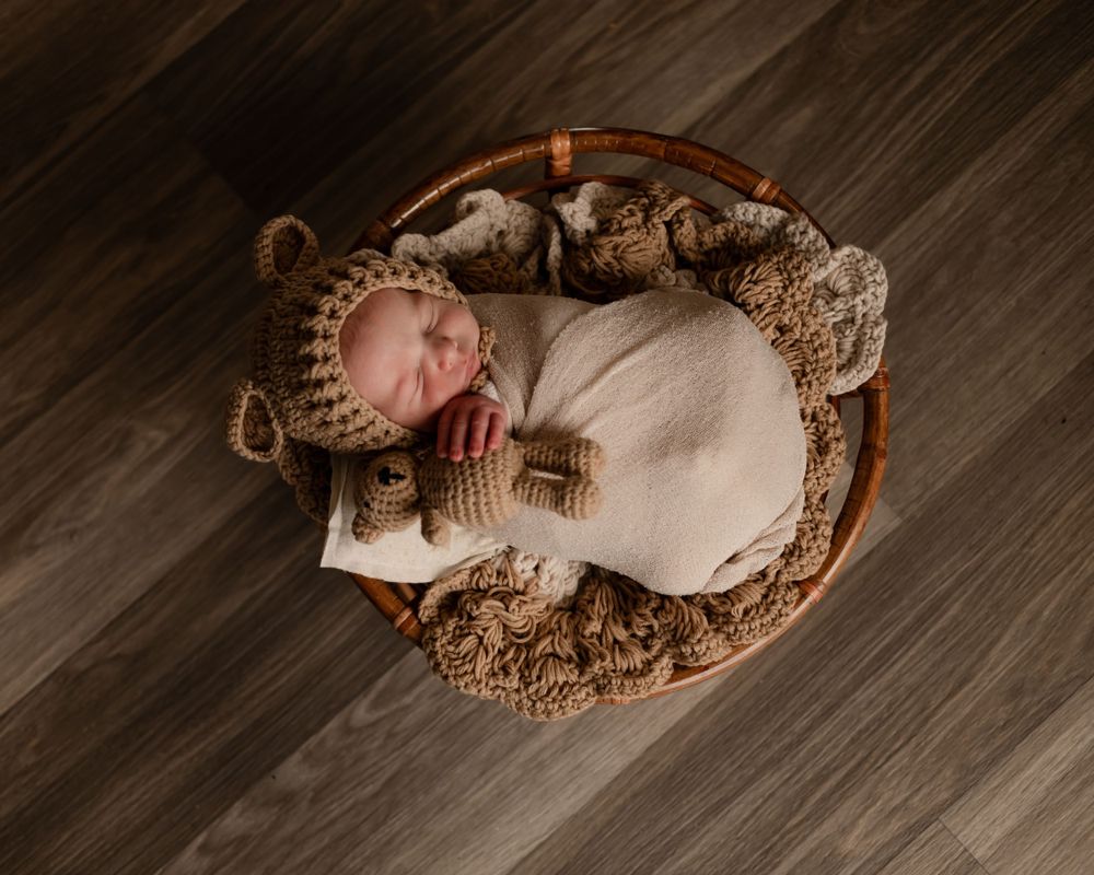 Sleeping newborn in knit hat resting in round wooden basket on dark floor.