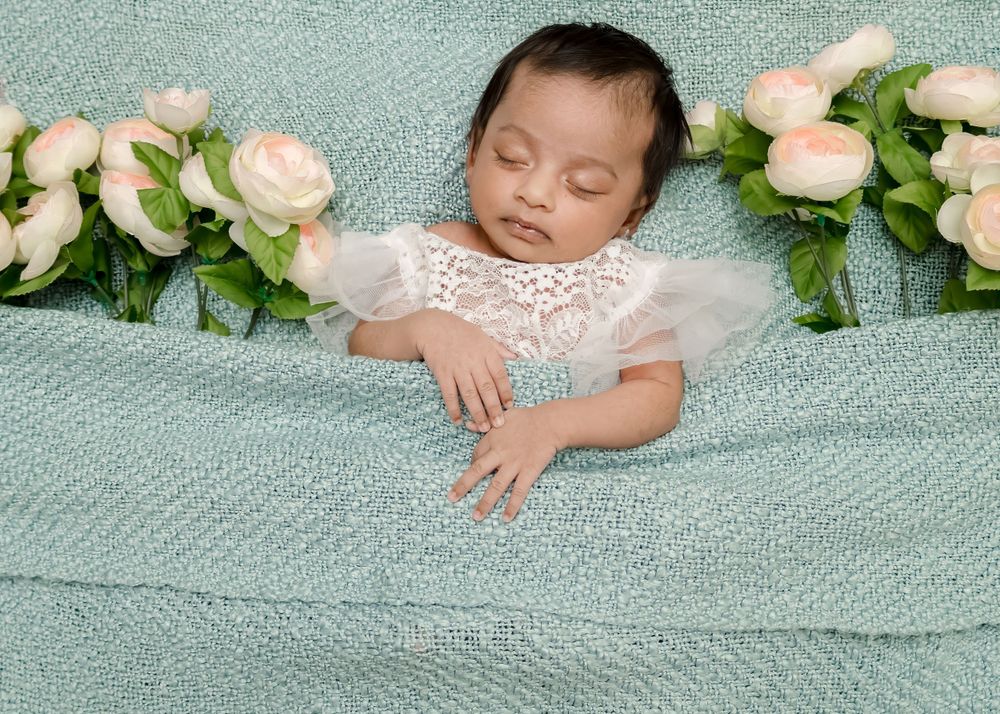 Sleeping infant in white lace dress surrounded by pink roses on mint green blanket.