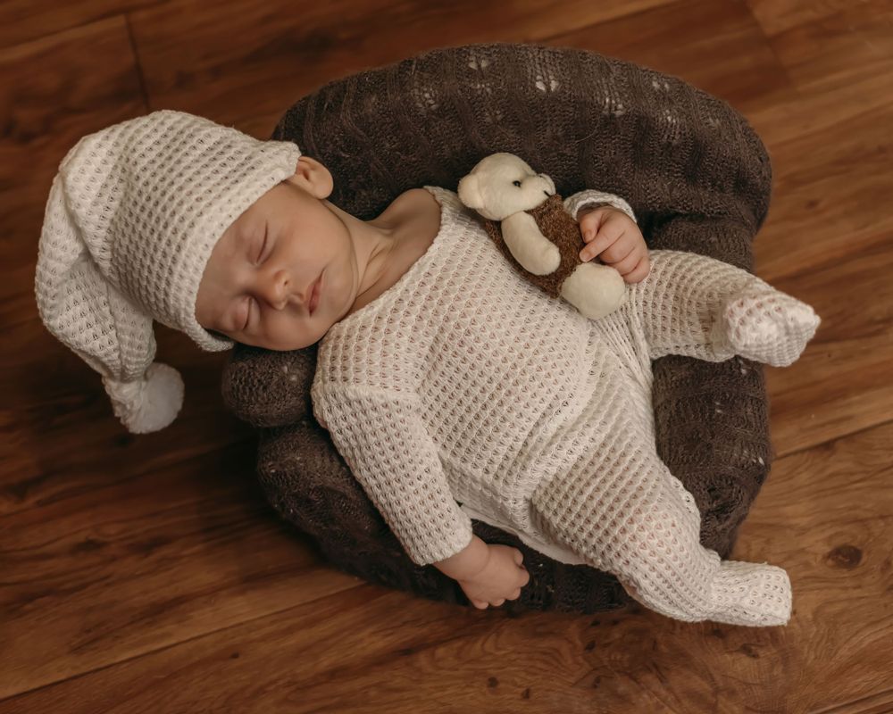 Sleeping newborn in white knit outfit laying on grey cushion with small plush toy.