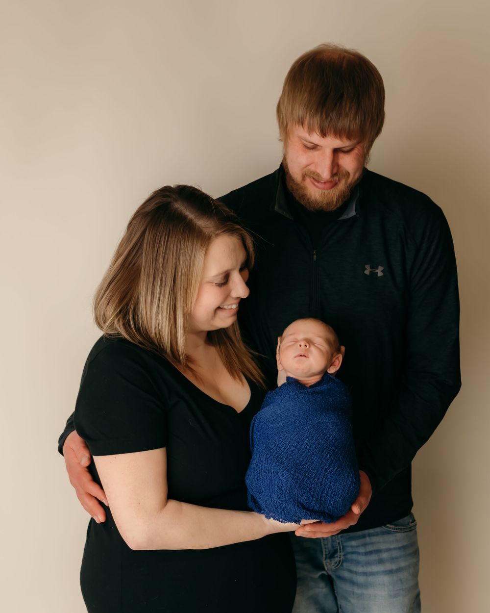 Family portrait with couple holding newborn wrapped in blue against neutral background.