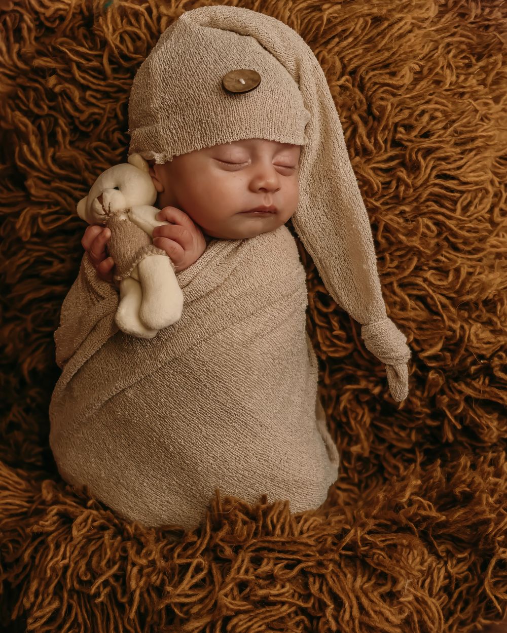 Sleeping newborn wrapped in beige fabric wearing bunny hat with plush toy on textured brown backdrop.