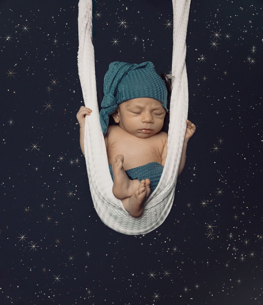 Infant sleeping peacefully in a white fabric hammock against a starry night sky background.