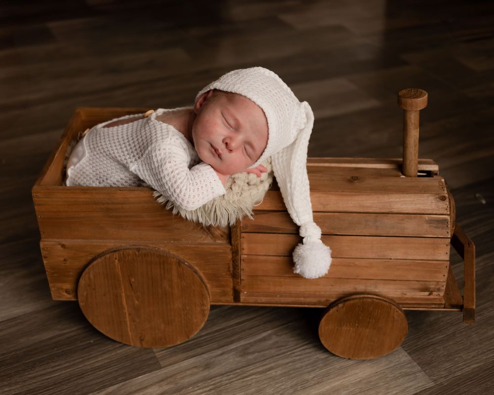 Sleeping baby in white outfit and hat lying in wooden toy truck prop.