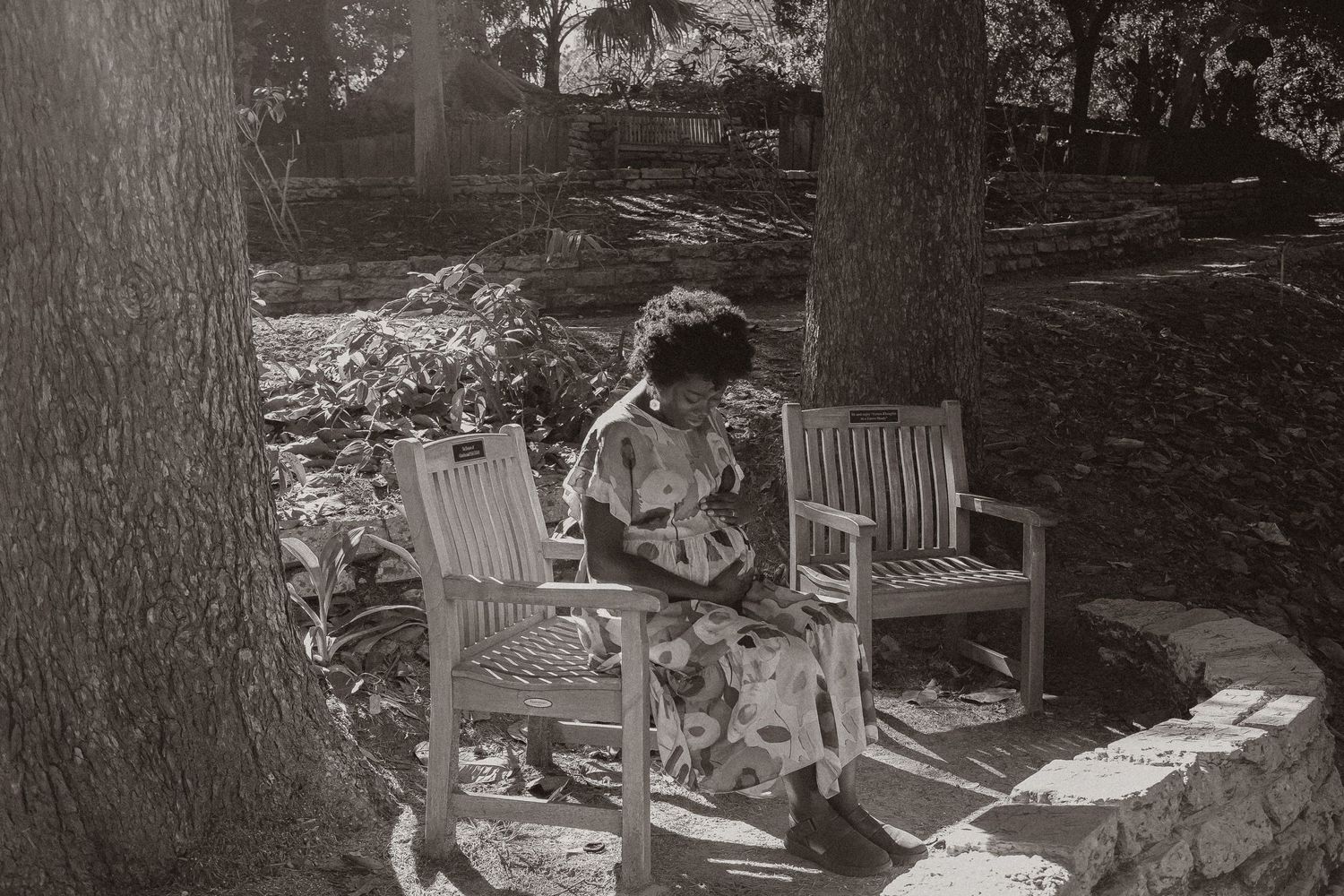 Vintage black and white photo of person seated in wooden chairs among garden trees.