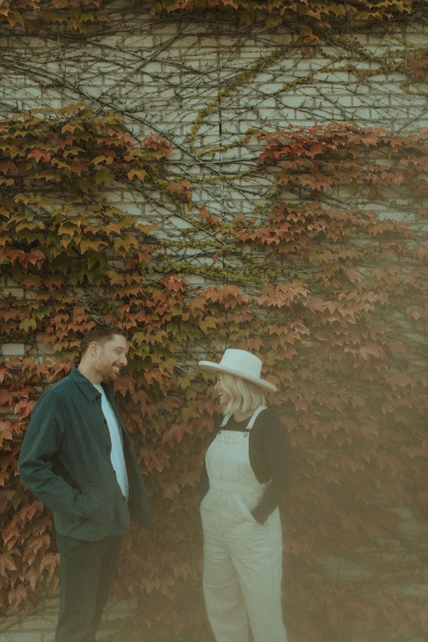 Two people stand together against a wall of changing fall foliage in stylish autumn outfits.