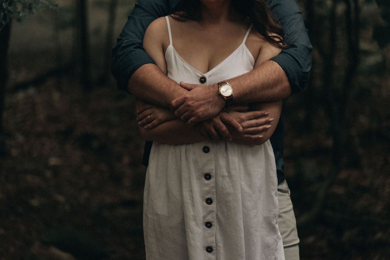 Intimate embrace captured from behind showing hands wrapped around in a forest setting.