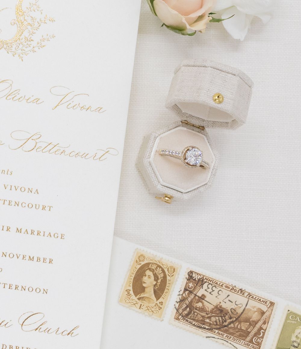 Wedding invitation details with gold calligraphy, vintage stamps and diamond ring in gray velvet box.