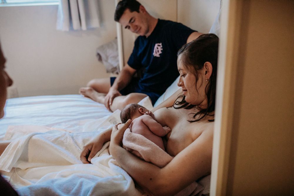 A tender moment at a home birth as a new parent holds a newborn baby while a midwife attends nearby and husband looks on lovingly.