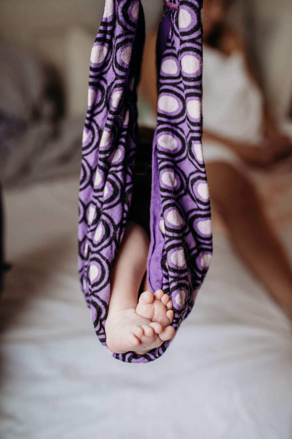 A tiny baby foot peeks out from a purple and black patterned fabric sling while getting weighed after home birth.