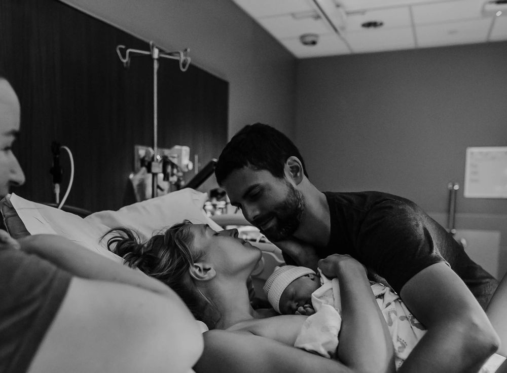Black and white photo of dad comforting mom right after birth as baby lays skin to skin on mama's chest.