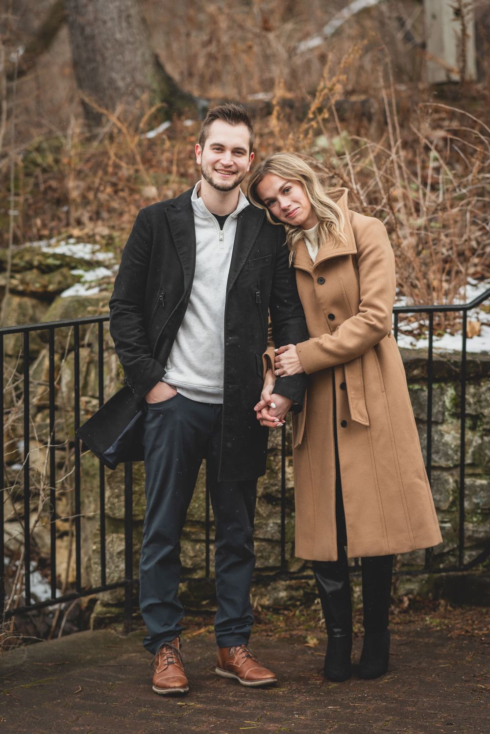 A couple in casual winter outfits posing by an iron fence in a snowy park setting.