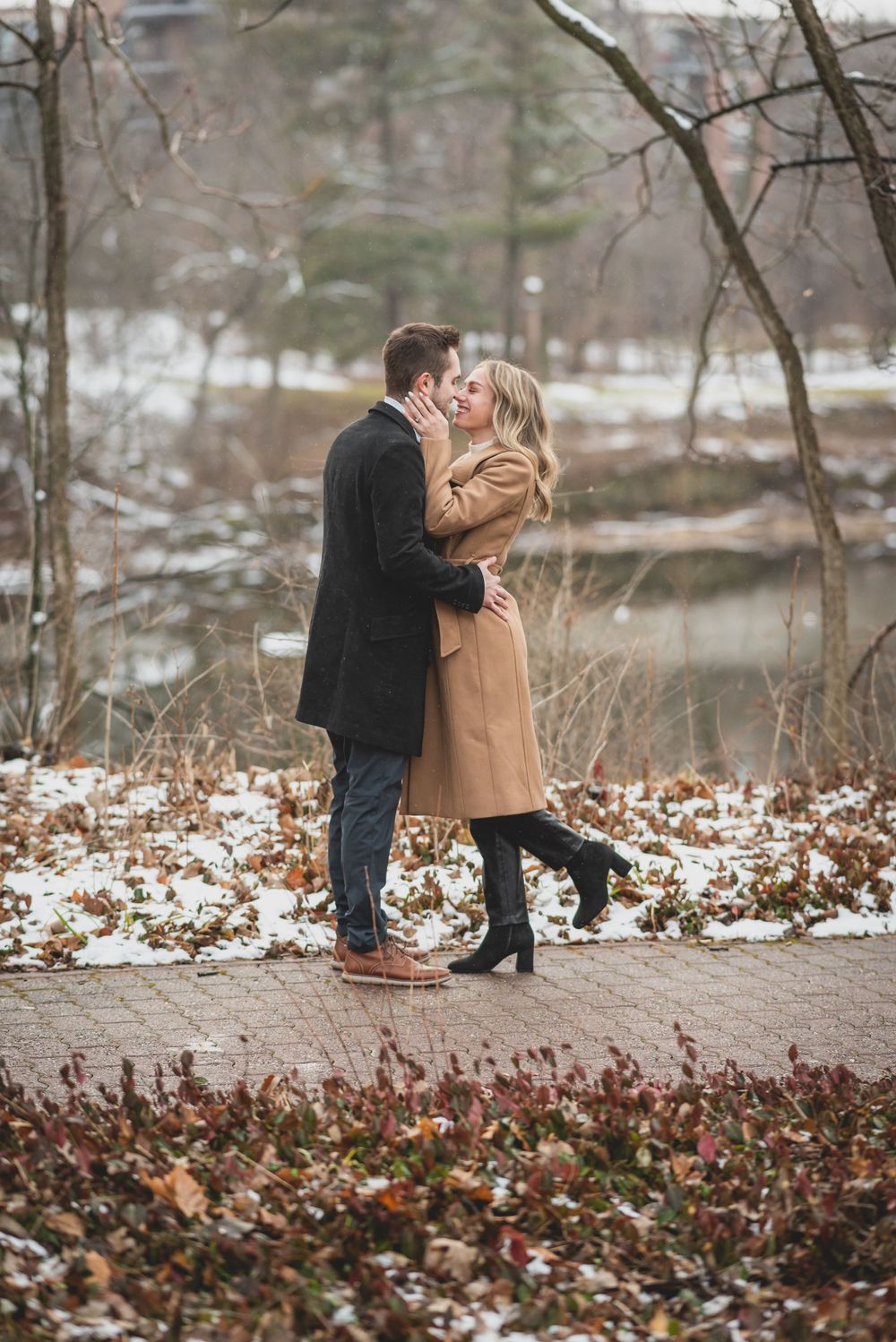 A couple walking and embracing on a snowy path surrounded by winter foliage.