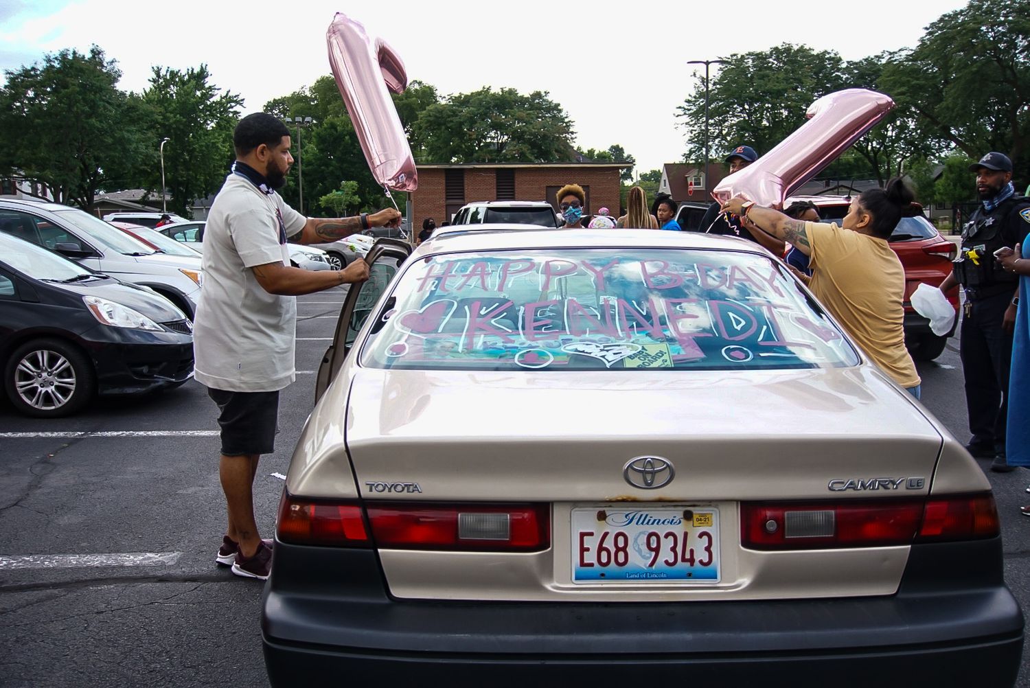 People decorating a white Toyota sedan with writing and balloons in a parking lot.