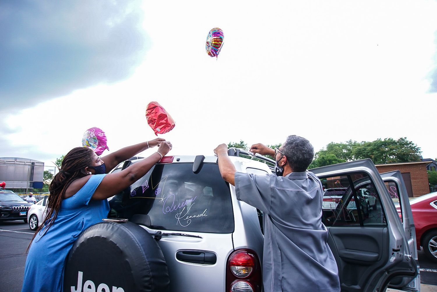 People reaching up to catch balloons floating above a parked Jeep.