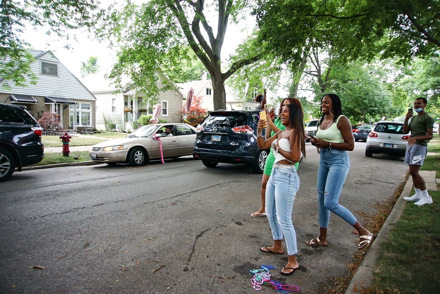 A group gathers outside in a residential neighborhood on a sunny day next to parked cars.