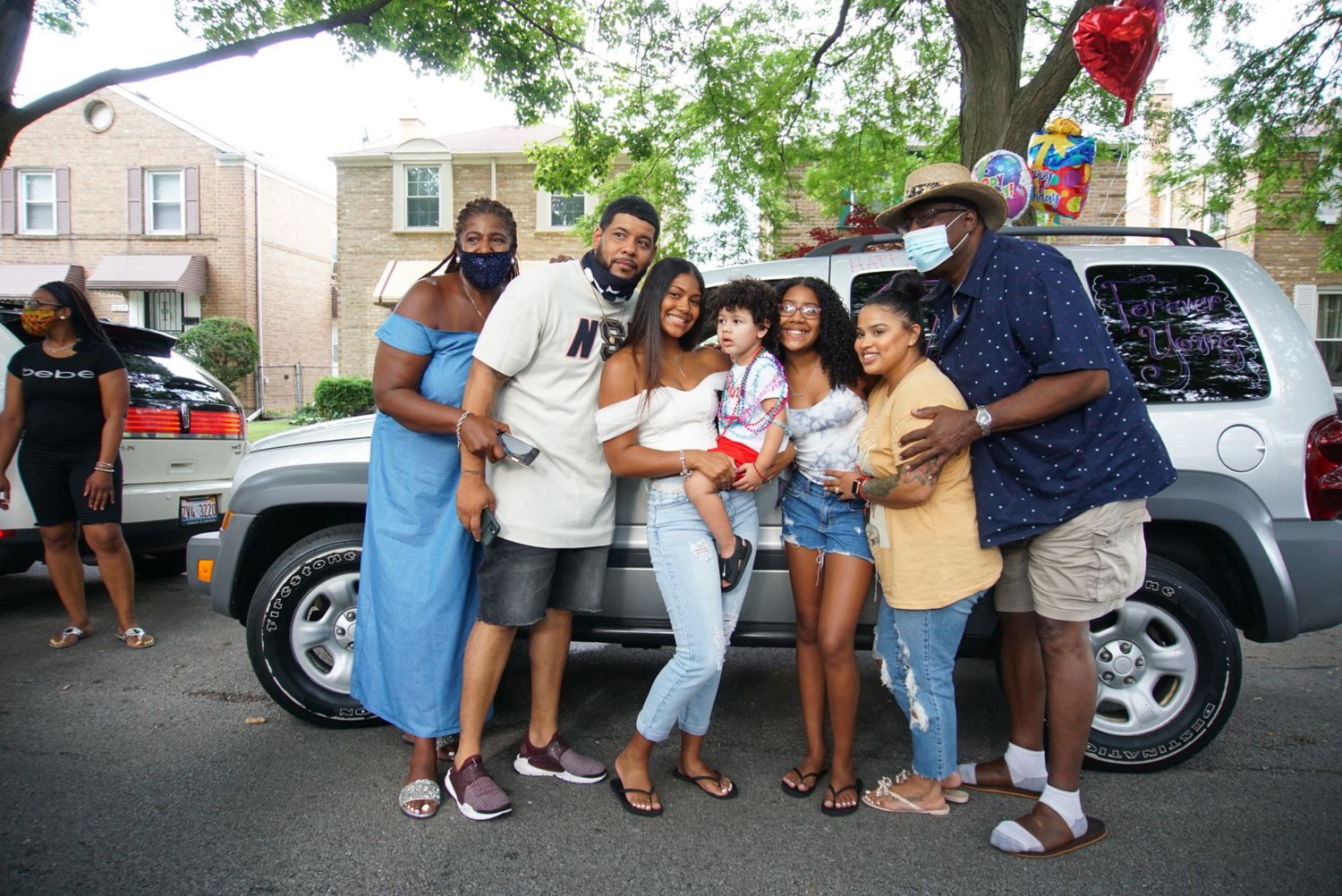 A group poses together in front of a silver SUV on a residential street with balloons visible.