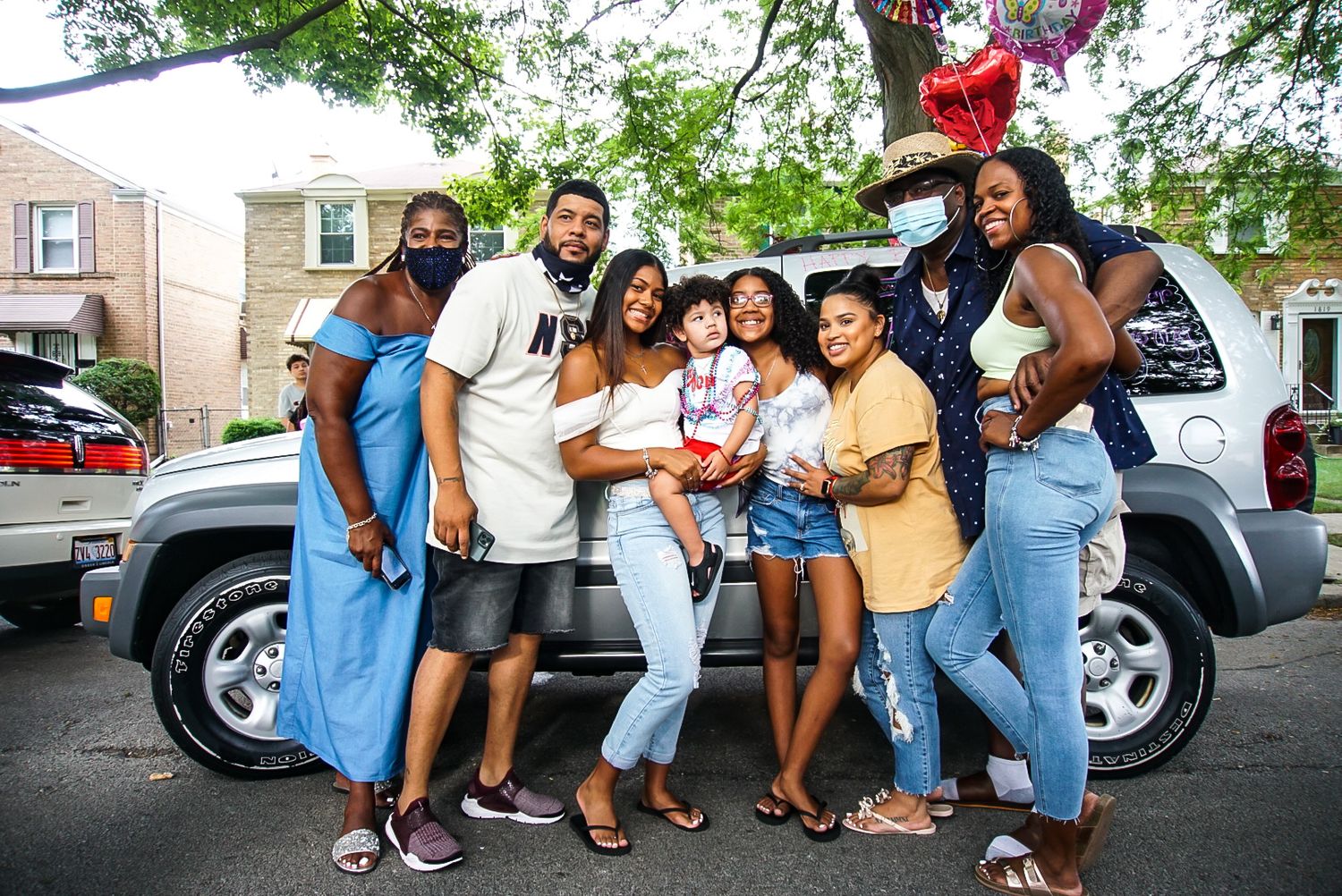 Friends gather for a celebratory photo by a parked SUV on a sunny street.