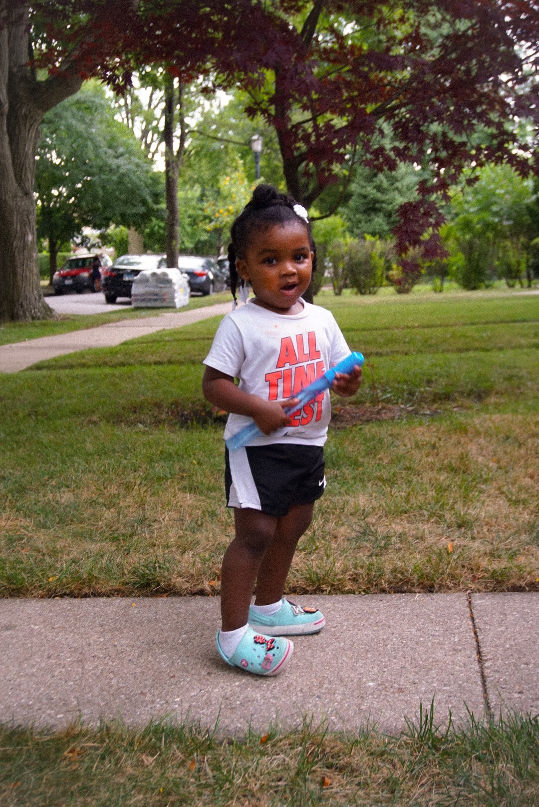 Small child in Nike shirt and shorts stands on suburban sidewalk wearing light blue shoes.