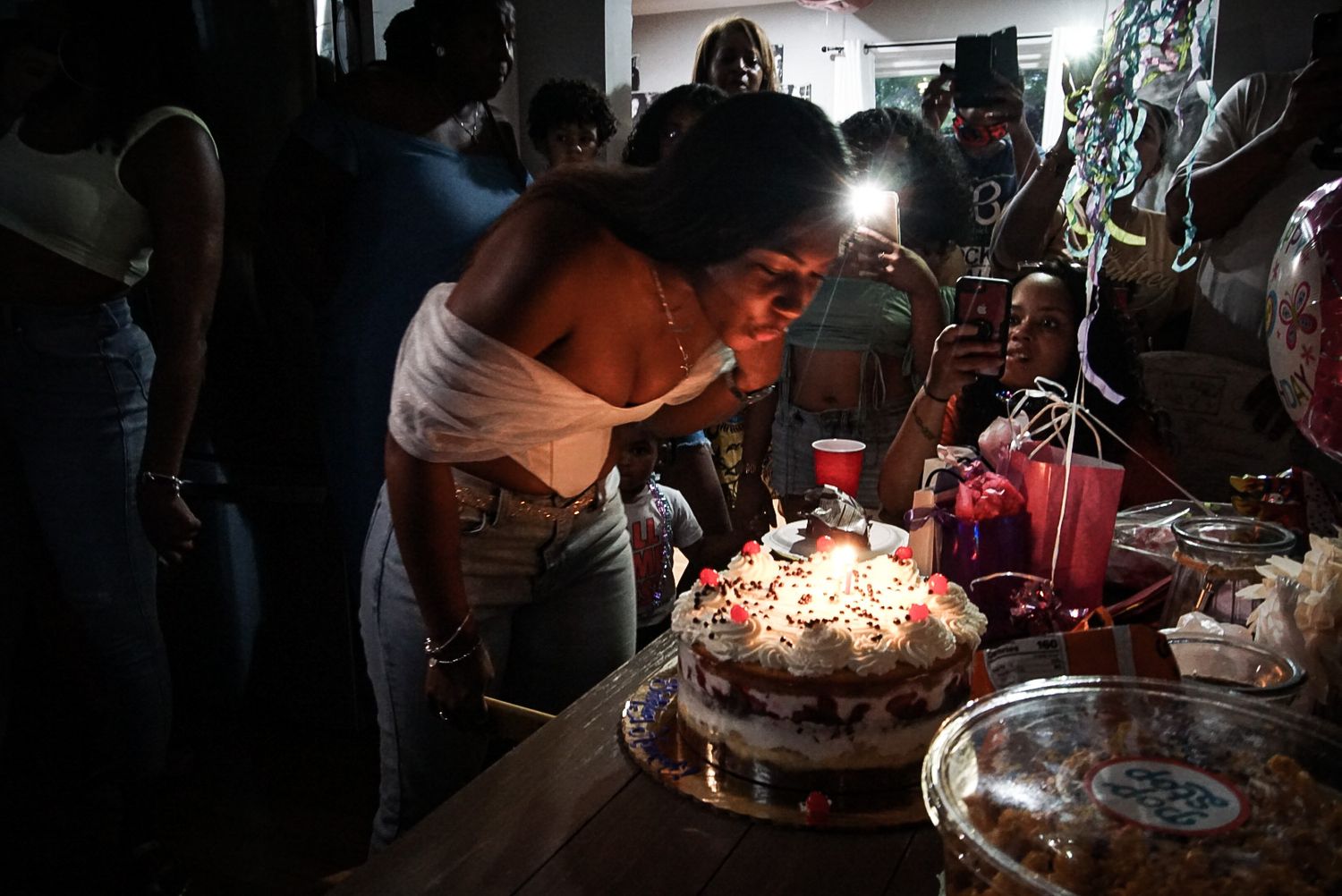 Person blowing out candles on a birthday cake in dark room surrounded by others.