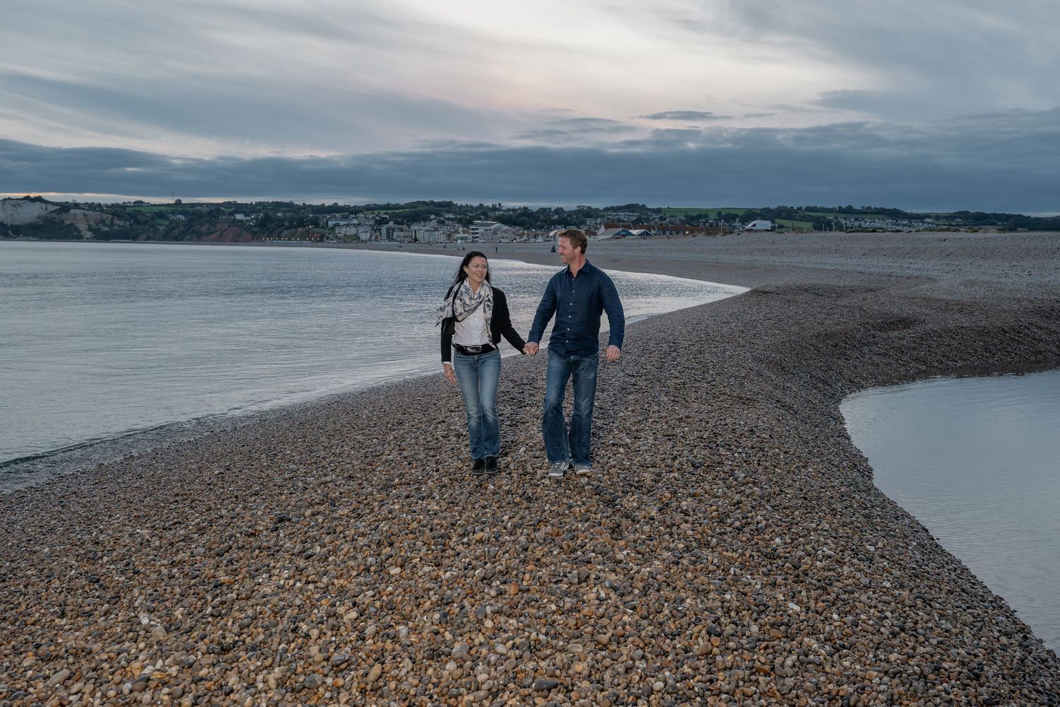 Couple walks hand in hand along rocky beach during sunset.
