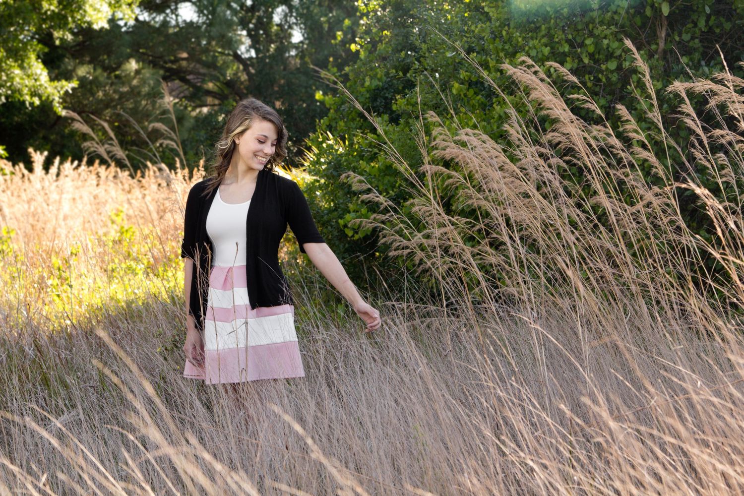 Teenage girl in pink and white dress with black sweater standing in a field of tall grass while the wind blows.