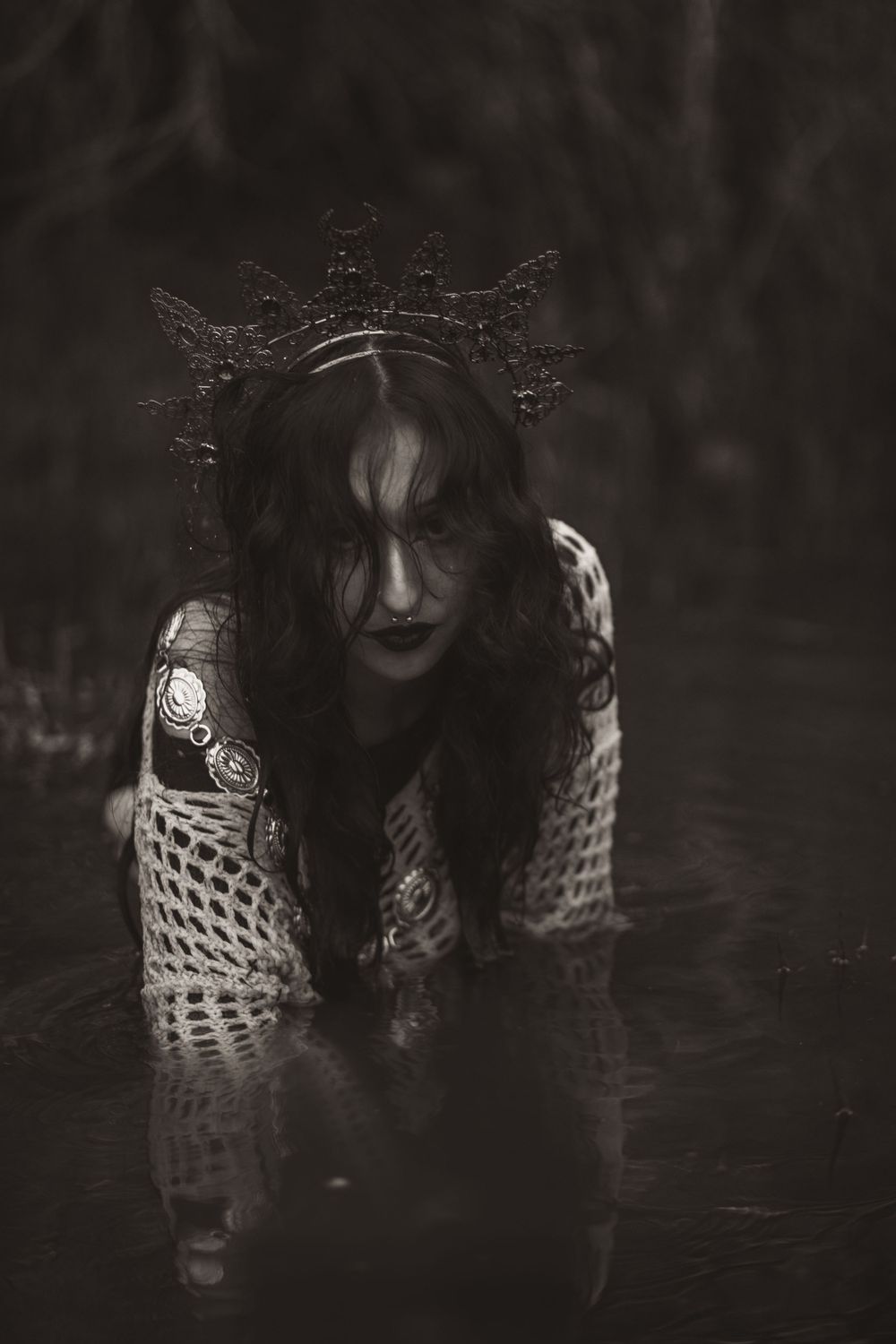 Ethereal dark figure in crown and mesh clothing crouches in atmospheric black and white portrait.