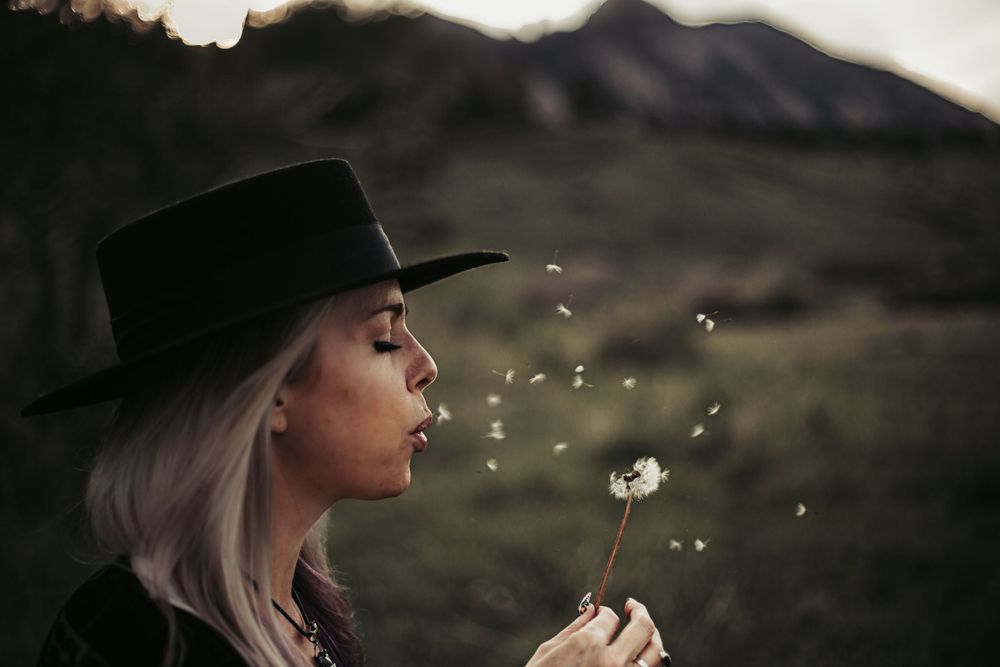 A dreamy portrait of someone blowing dandelion seeds at sunset with mountains in the background.