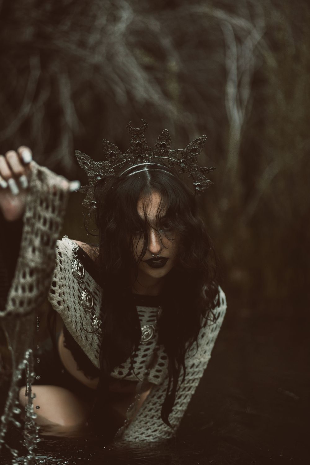Dark gothic figure wearing an ornate crown and lace dress emerges from shadows in moody forest setting.