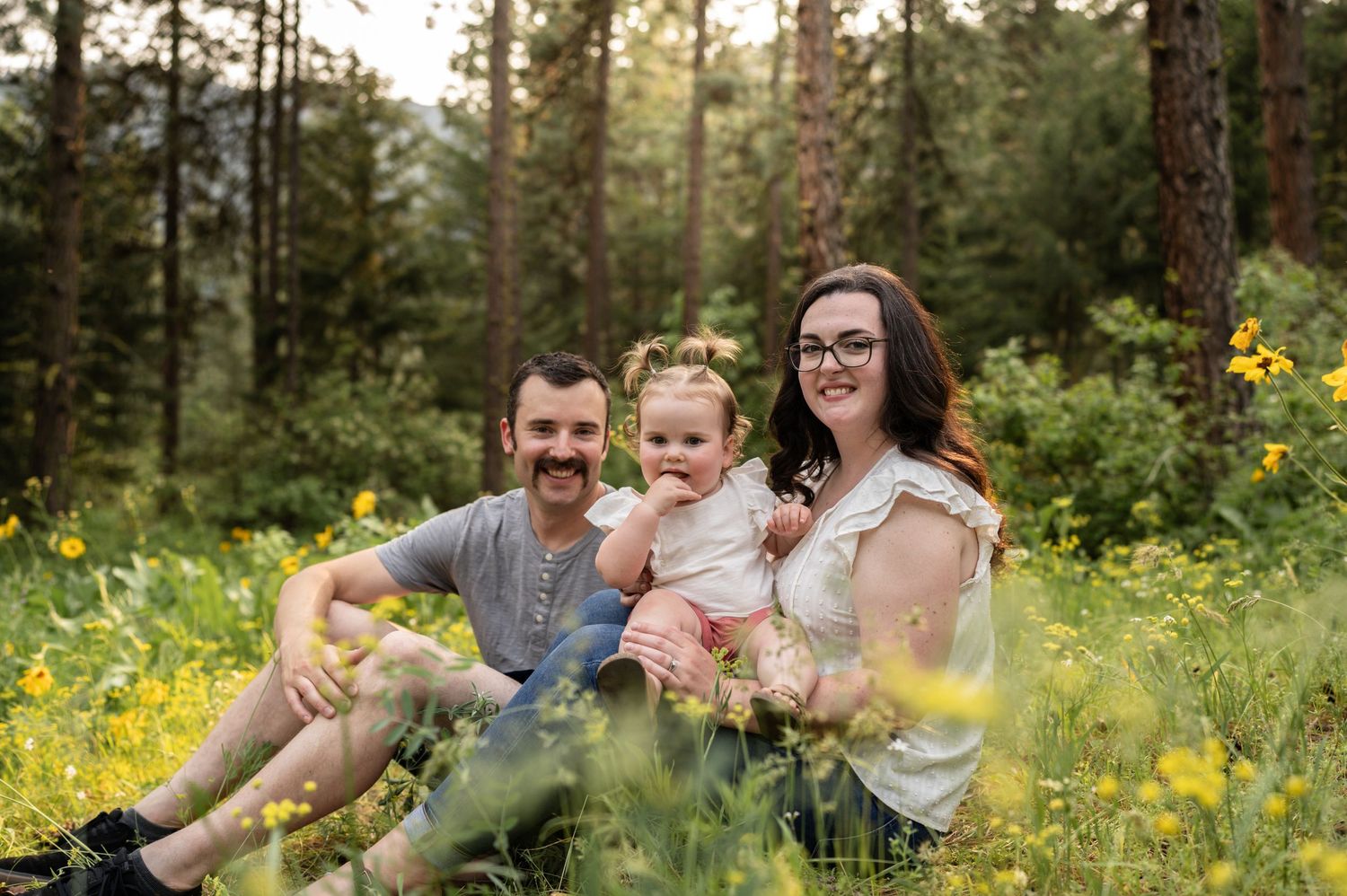 A loving group sits together in a meadow filled with yellow wildflowers and pine trees.
