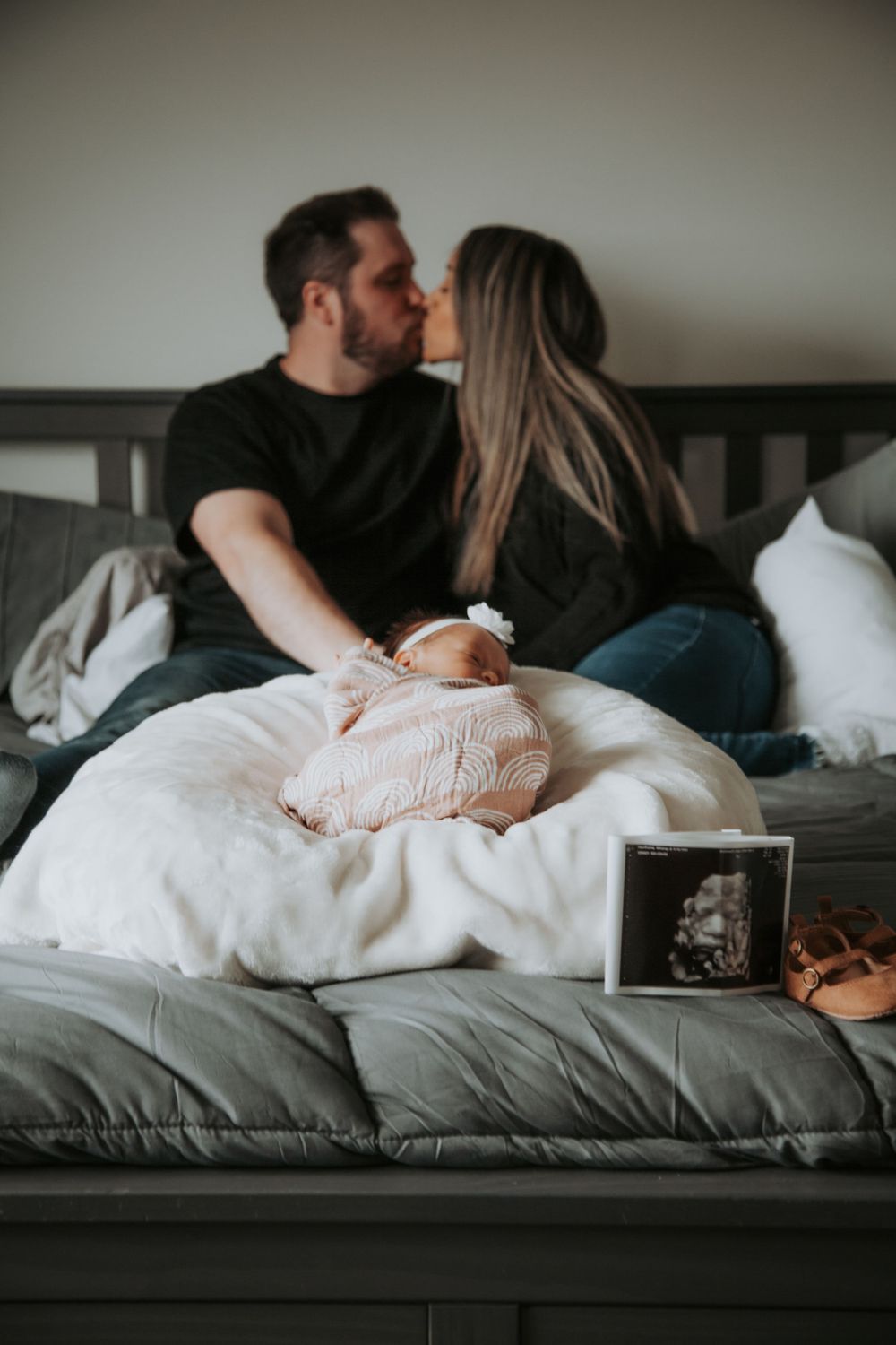 Couple kissing on a bed behind their swaddled newborn baby, with an ultrasound photo and baby sandals nearby in a newborn photo session.