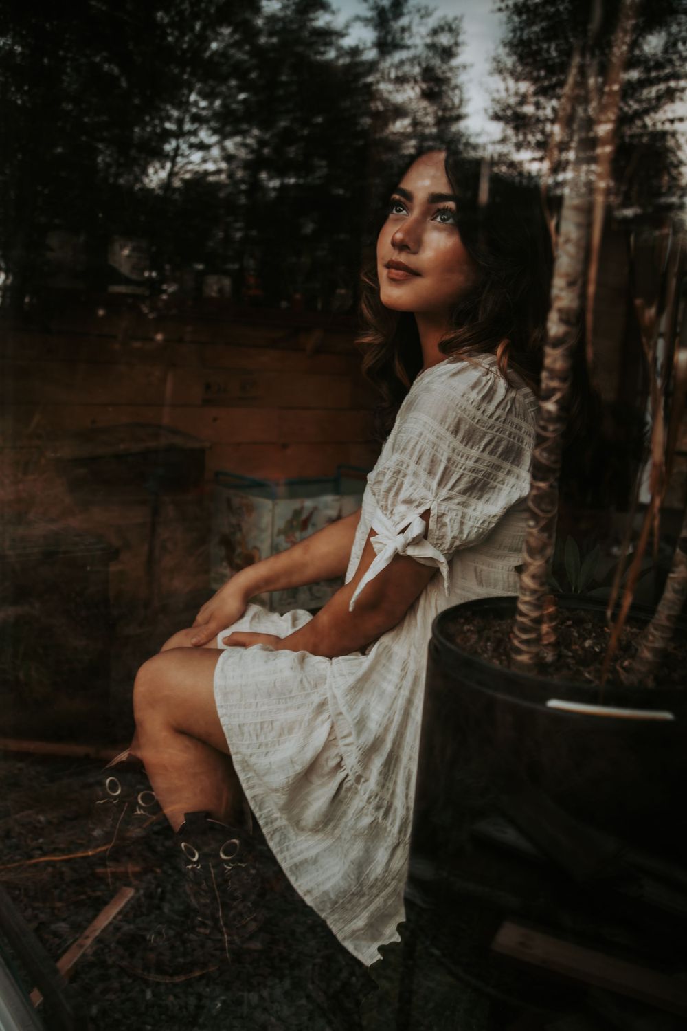 A woman in a white dress and boots sits beside a potted tree while a window reflection of forest trees overlays the moody portrait.