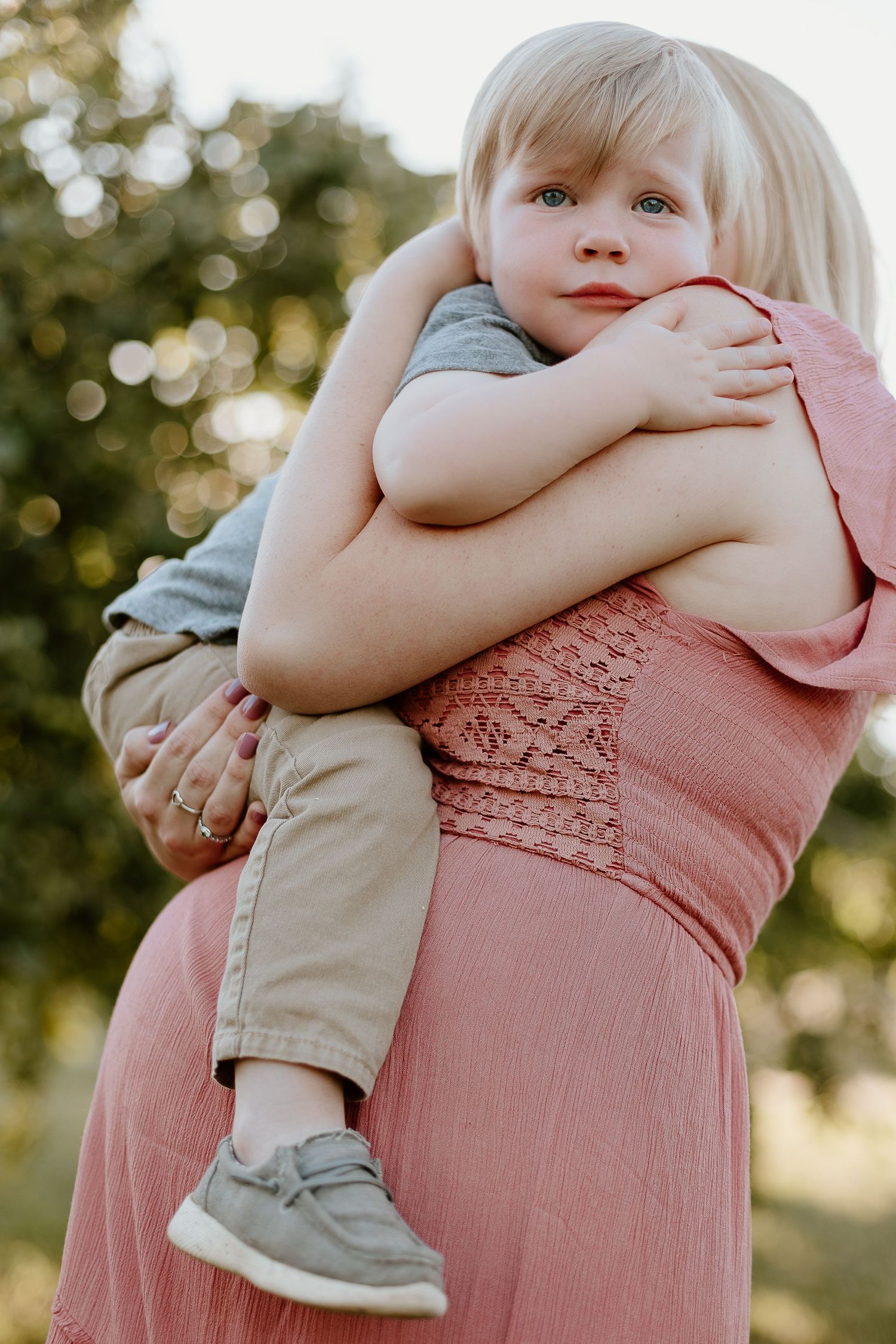 A small child in grey being held and hugged by their mother wearing a pink dress outdoors during maternity session.