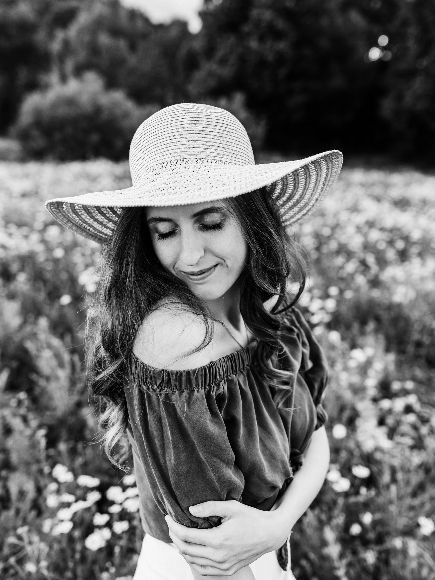 A dreamy black and white portrait of a woman in a wide-brimmed sun hat standing in a wildflower field.