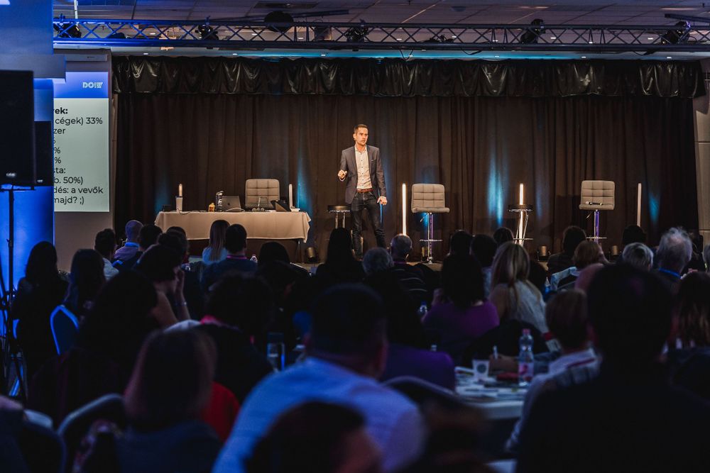 Conference speaker presenting on stage to seated audience during evening business event.