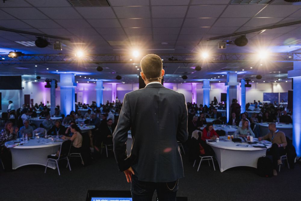 Speaker addressing large audience in formal event space with blue ambient lighting.