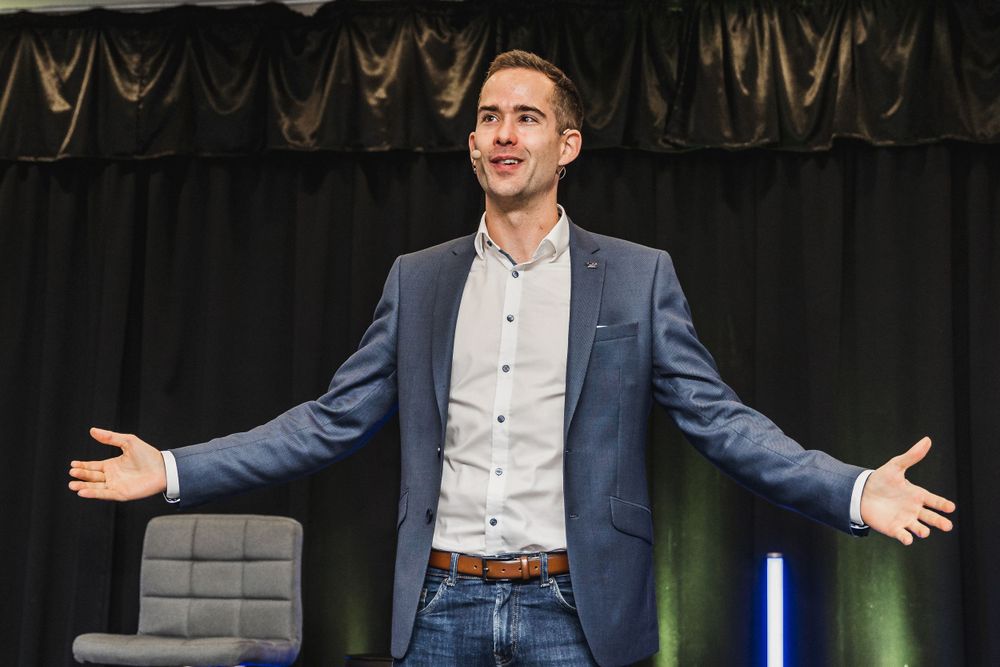 Speaker on stage wearing business casual attire with arms outstretched.