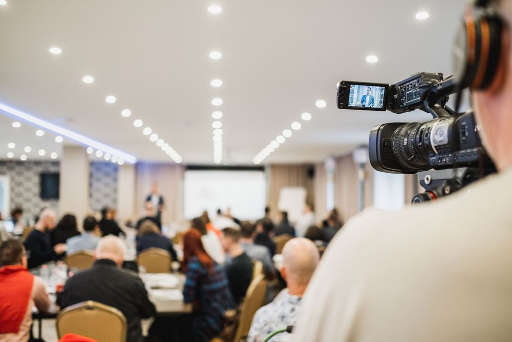 Professional video camera recording conference presentation from back of crowded room.