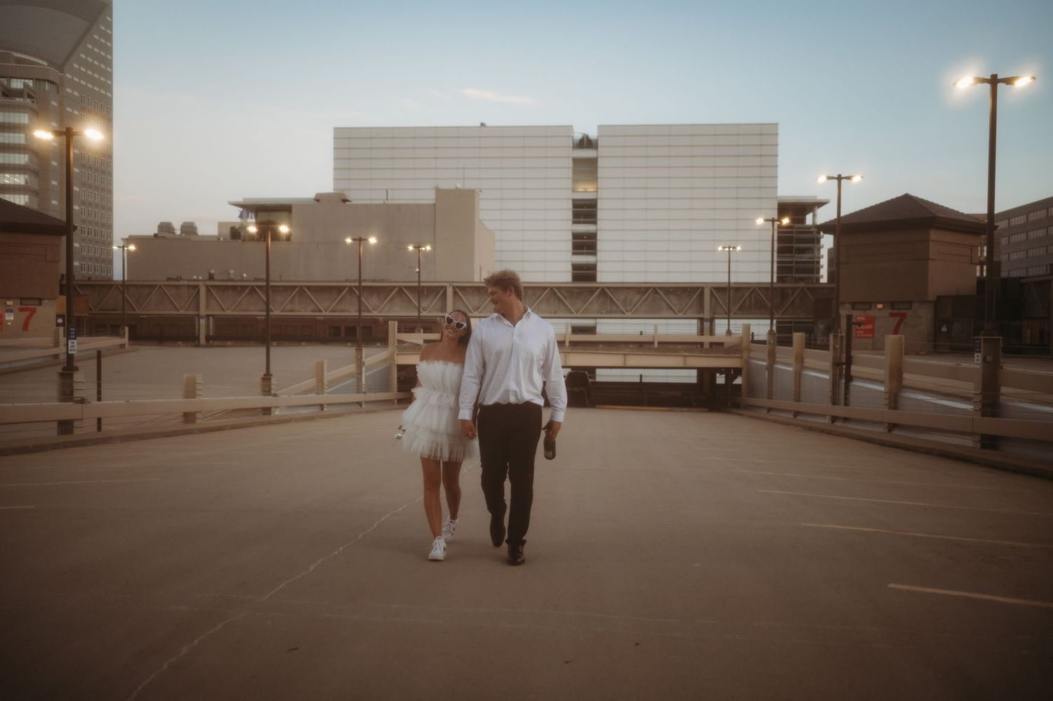 Couple walking together at sunset in an empty parking lot with a large building in the background.