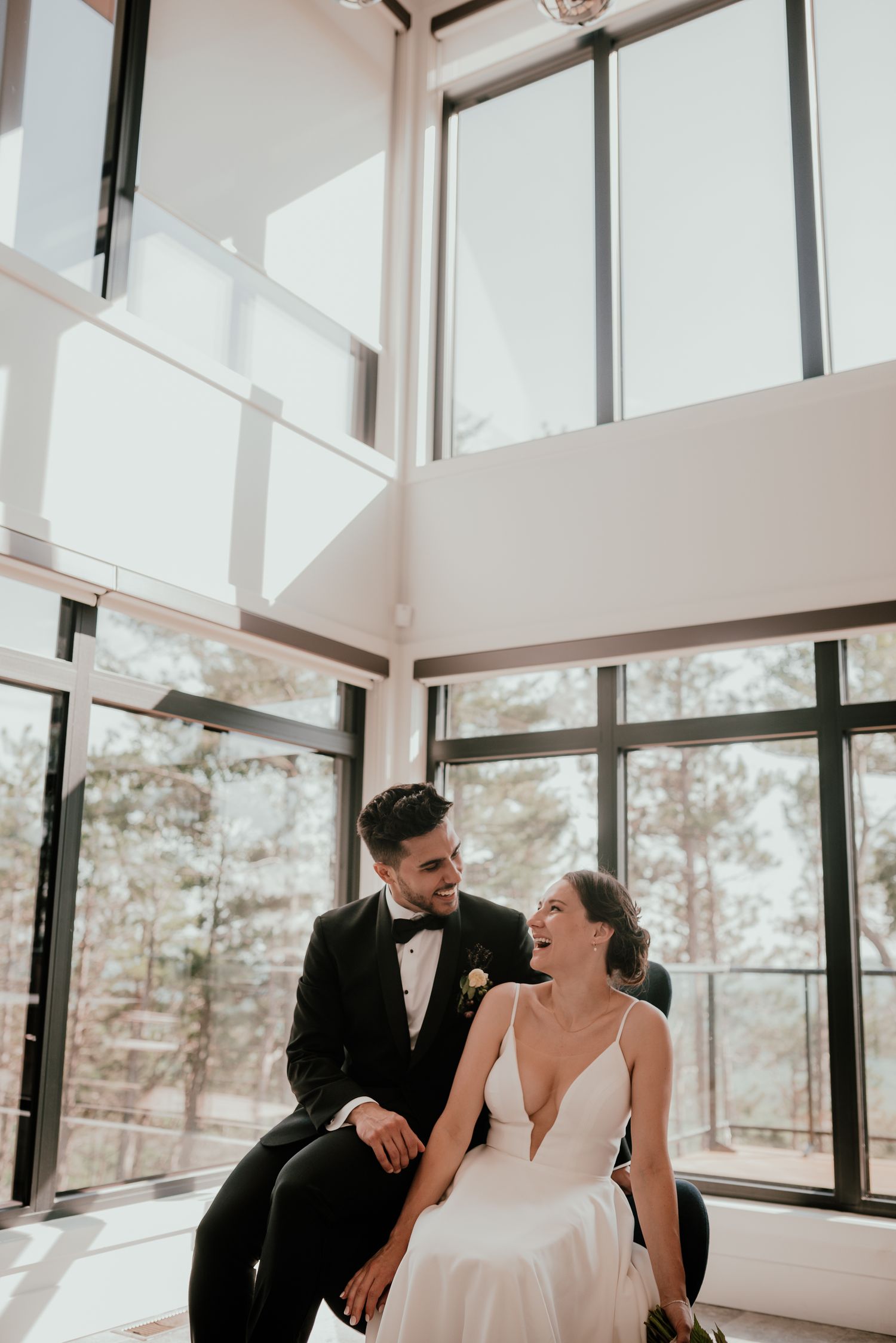 A couple shares a joyful moment together by large windows in a bright, modern room during their wedding day.