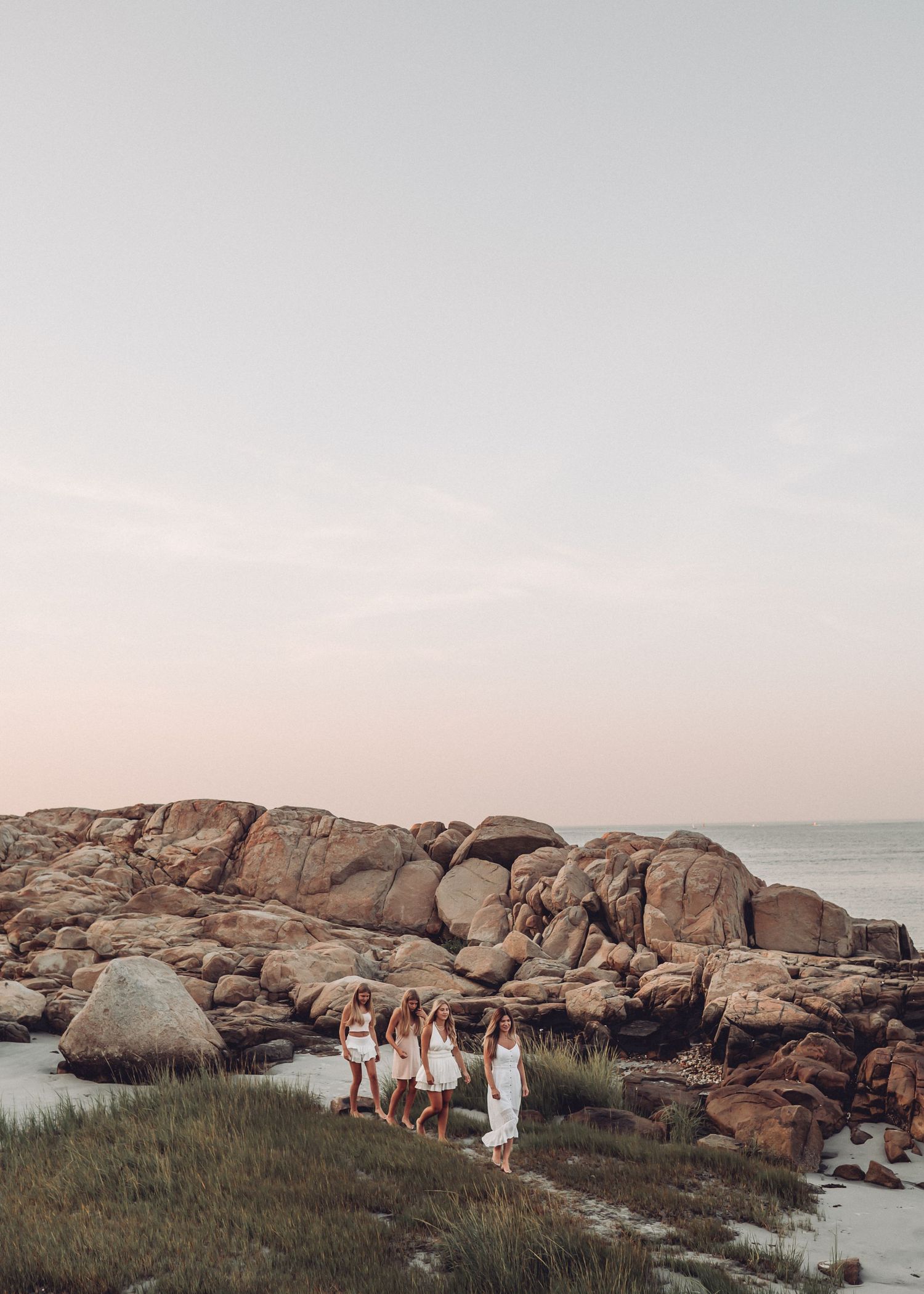 Group in white dresses standing on rocky coastal landscape at sunset.