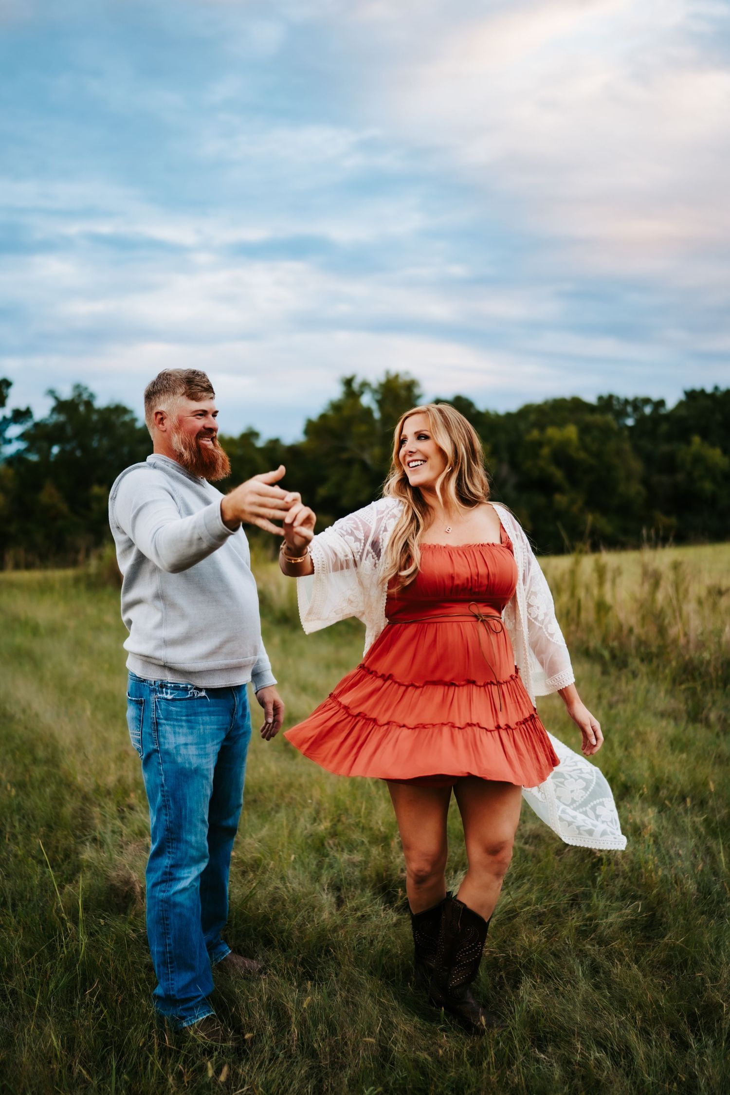 A couple dances in a field at sunset, the woman wearing a red dress and the man in casual attire.