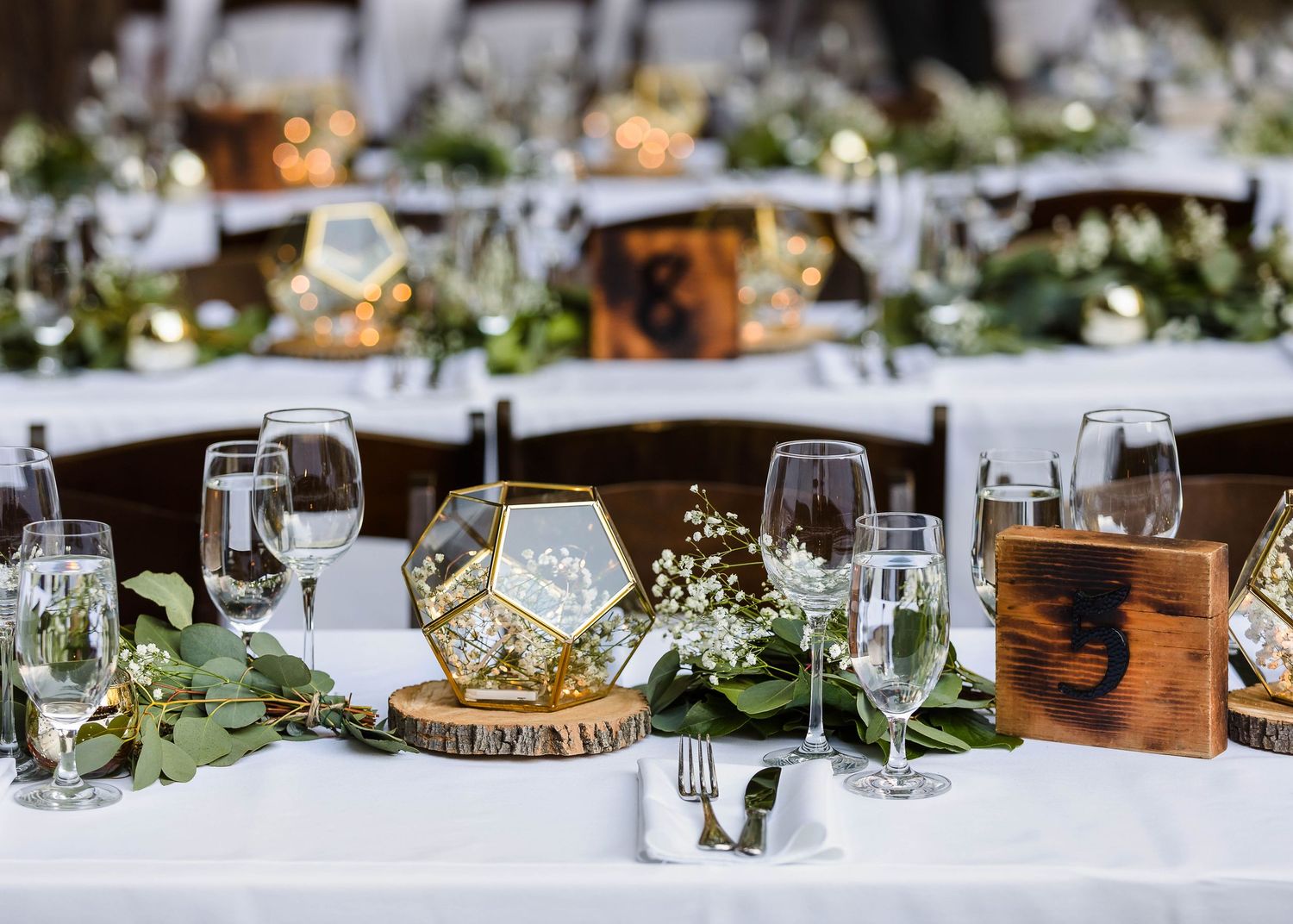 Elegant table settings with wooden table numbers, geometric centerpieces, and greenery at an outdoor reception.
